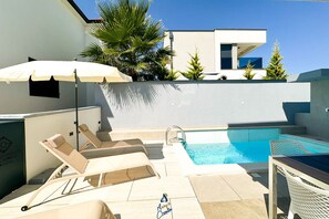 Ein Poolbereich mit Liegestühlen, Sonnenschirm und einem modernen Gebäude im Hintergrund.