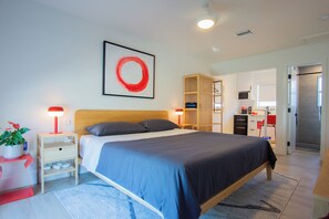 A modern bedroom with a bed, bedside tables, a red lamp, a framed artwork, a wooden wardrobe, and a kitchenette area. - Modern 2BR/2BA • Pool • Parking • Near Beach / Victoria Park / Las Olas (Fort Lauderdale)