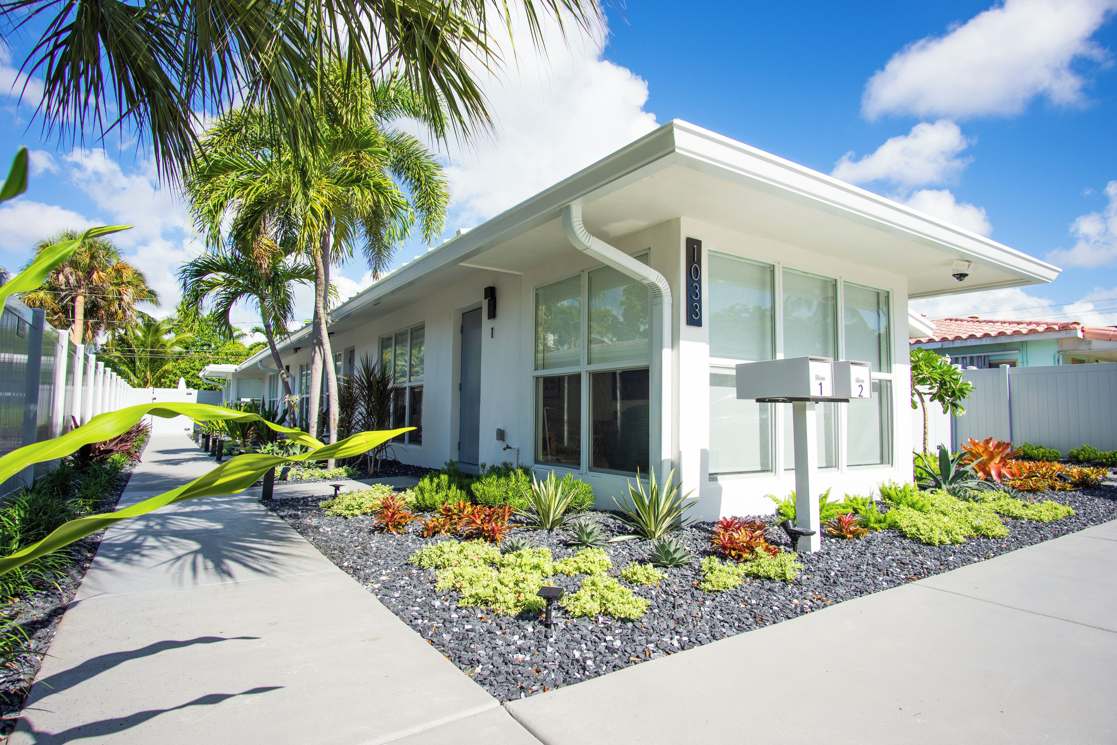 A modern two-story house with a flat roof, large windows, and a well-maintained garden with various plants and a pathway.