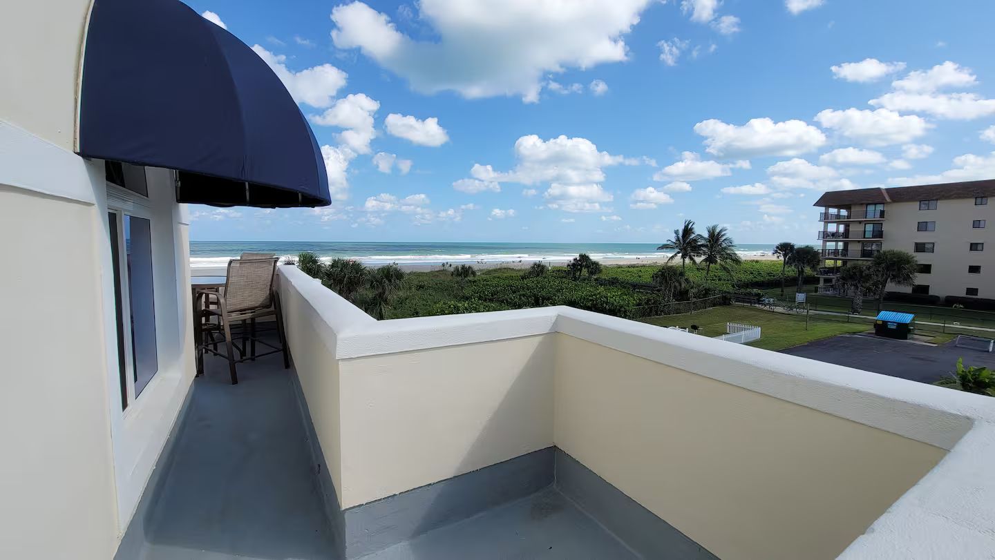 Ein Balkon mit Blick auf Strand und Meer, ein Stuhl und ein Teilansicht eines Gebäudes.