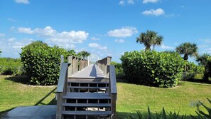 Un escalier en bois mène à une entrée fermée par un portail, qui est elle-même entourée d’une pelouse et d’arbustes bien entretenus.