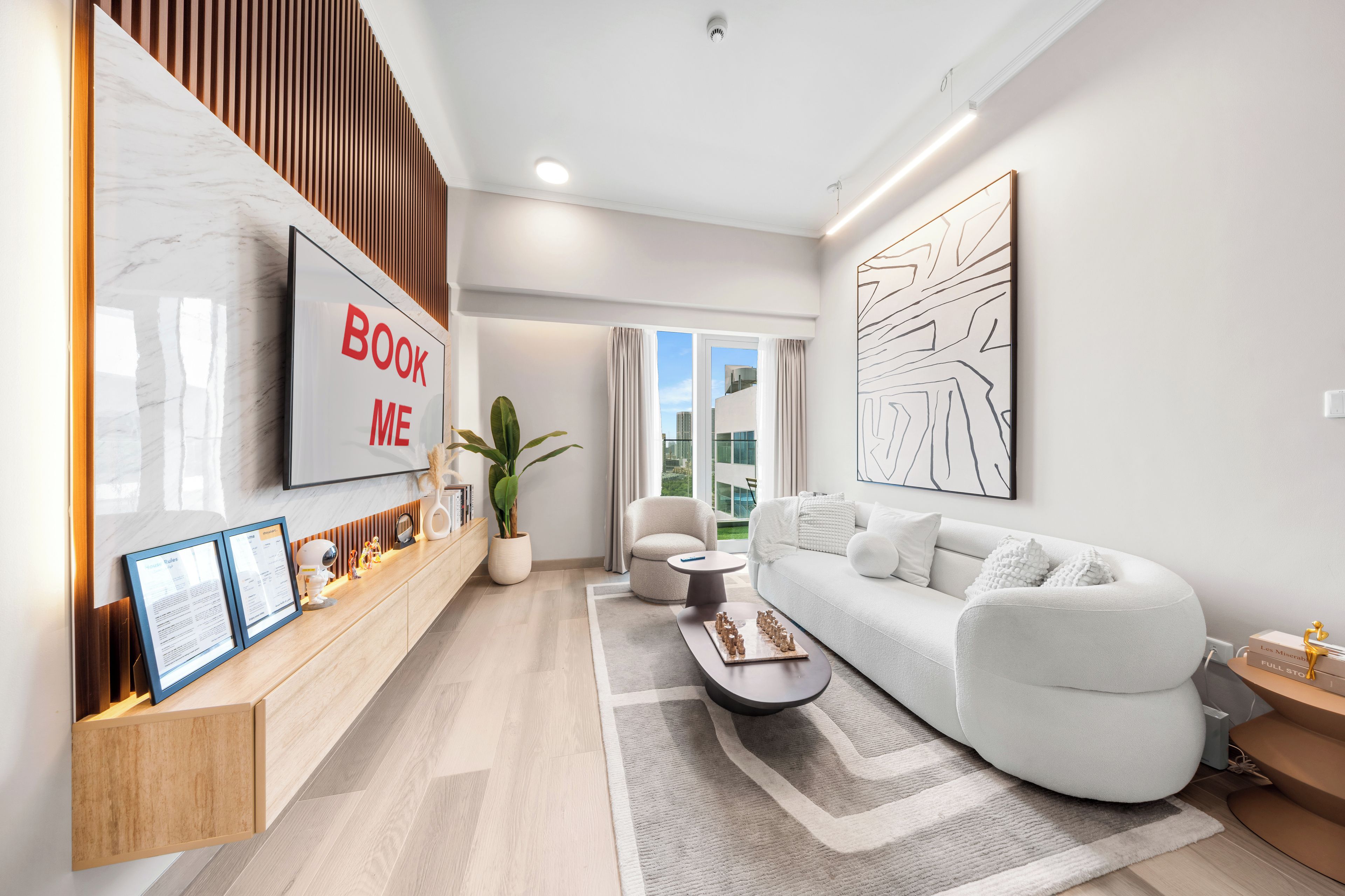 A modern living room with a sofa, a coffee table, a plant, and a 'BOOK ME' sign on the wall.