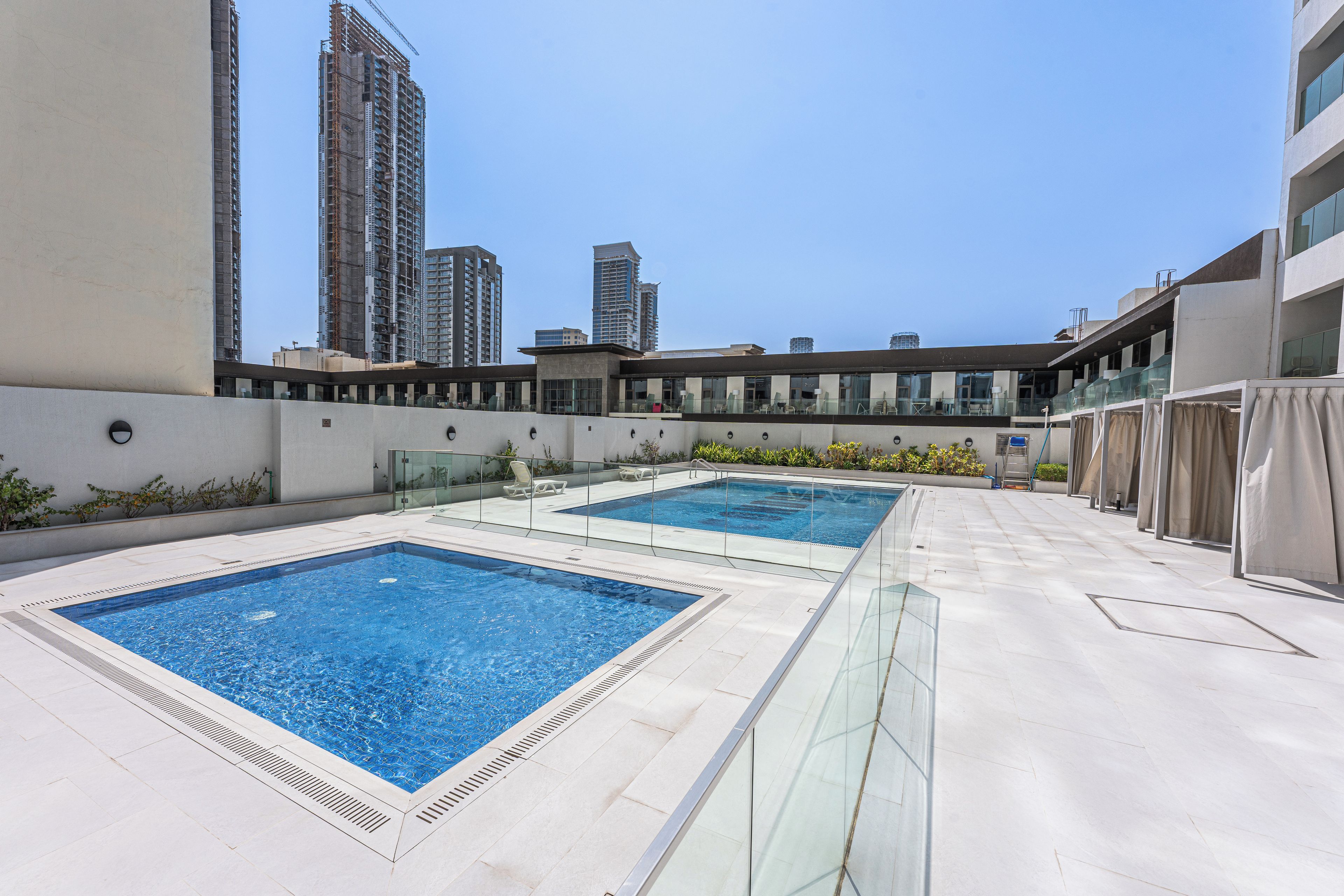 Un espace piscine sur le toit, comprenant deux piscines, entouré d’un bâtiment moderne doté de grandes fenêtres et offrant une vue sur les gratte-ciel en arrière-plan.