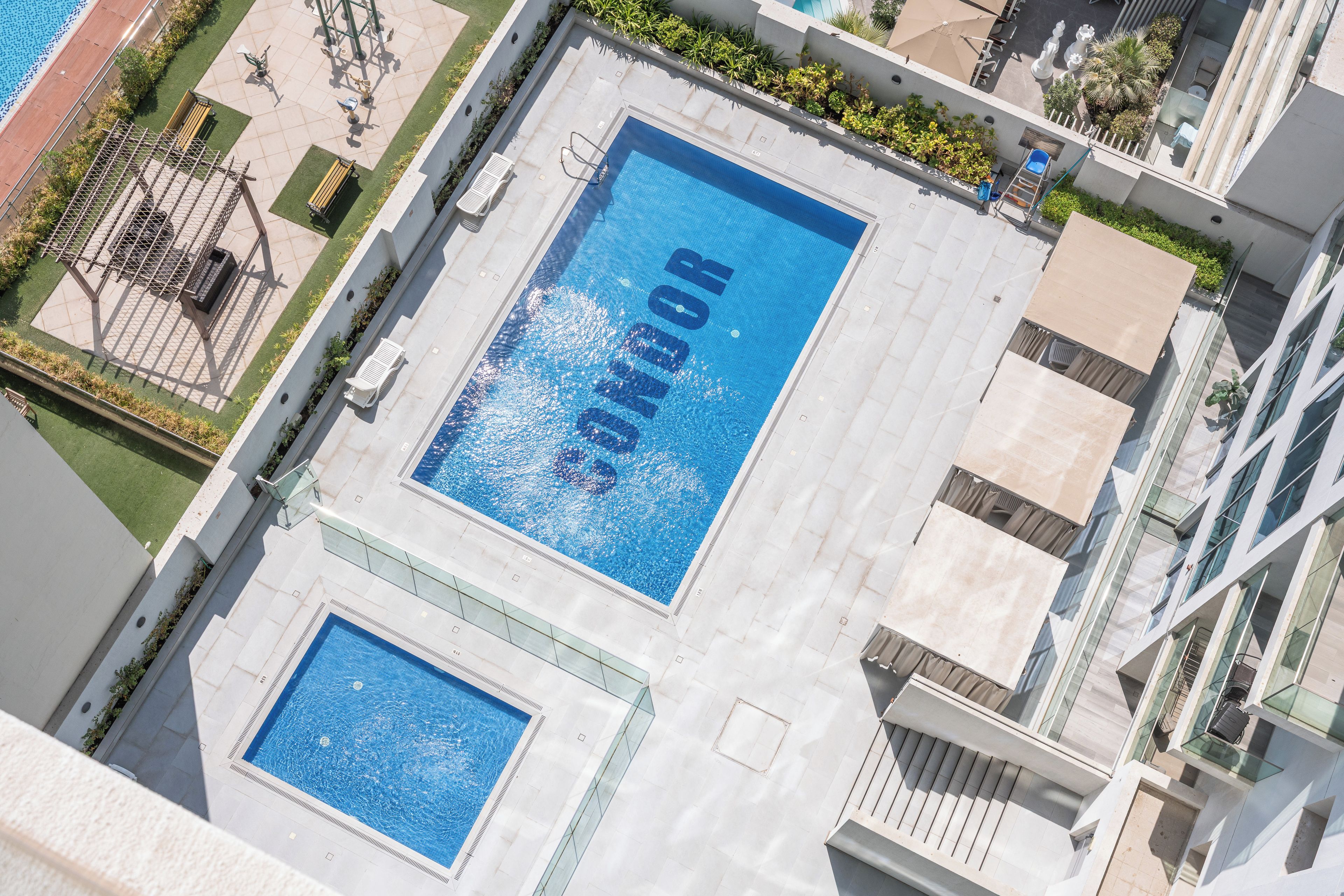 A rooftop swimming pool with a clear blue water surface, surrounded by a white tiled deck and a low green lawn.