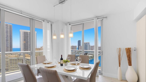 A modern dining area with a white table set for four, large windows, and a city view. - Beachwalk Resort Style 3BR Comfort Resort Vibes (Hallandale Beach)