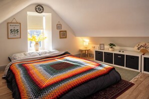 A cozy bedroom with a sloped ceiling, a colorful knitted blanket, a bedside table with a lamp, and a window with a view of the outdoors.