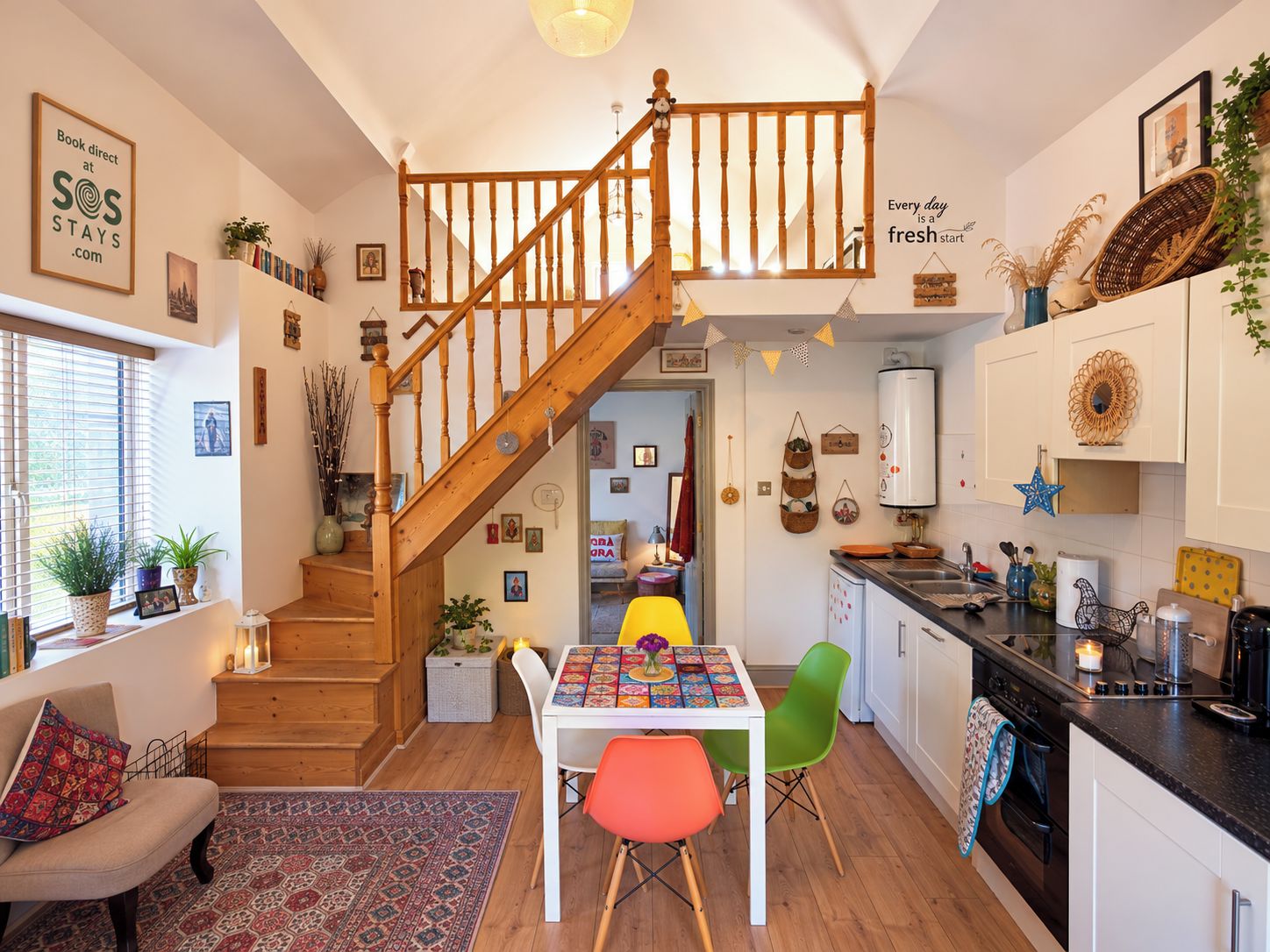 A cozy kitchen with a wooden staircase, a dining table with colorful chairs, and various kitchen appliances.