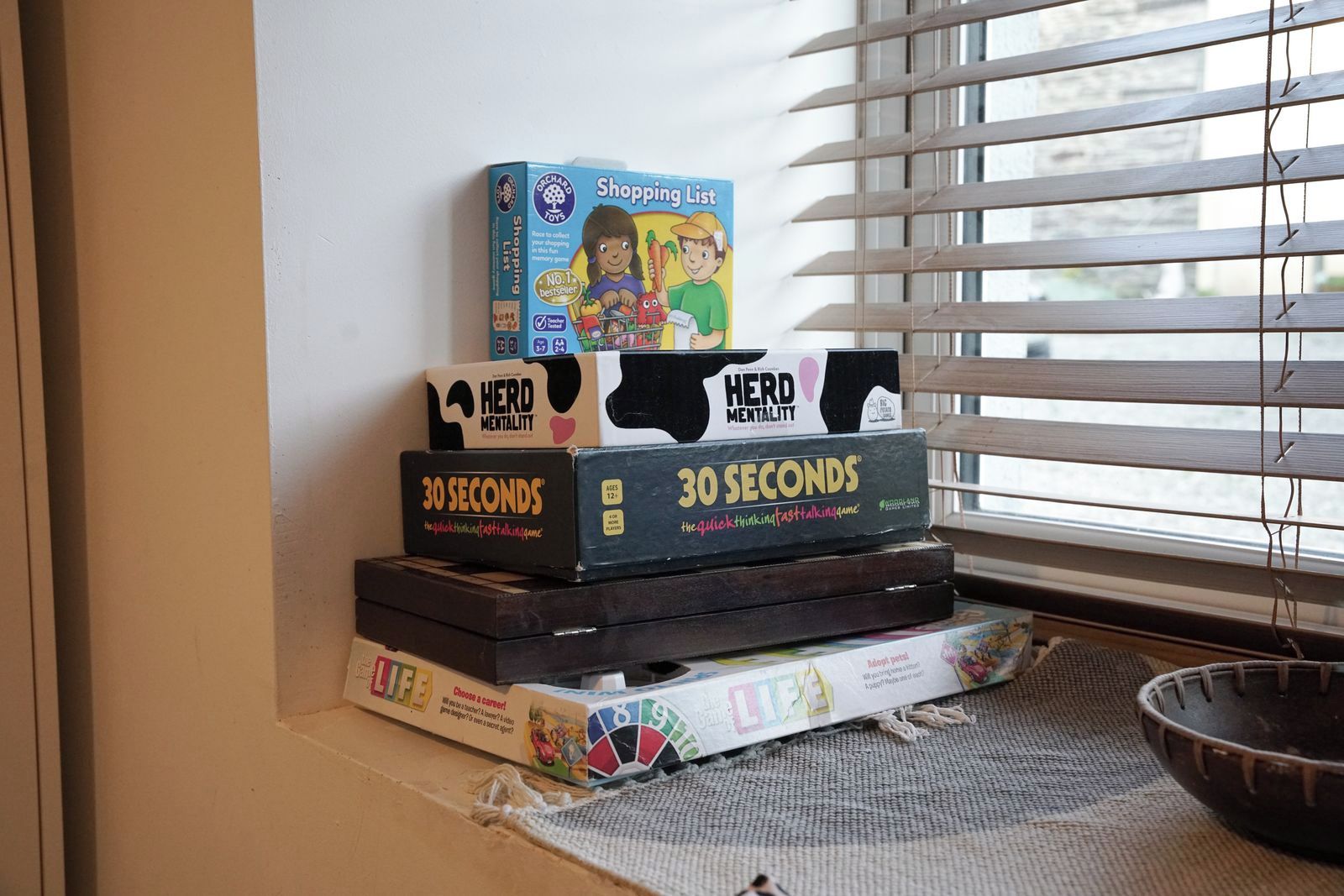 A stack of board games on a windowsill with blinds partially closed.