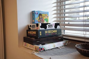 A stack of board games on a windowsill with blinds partially closed.