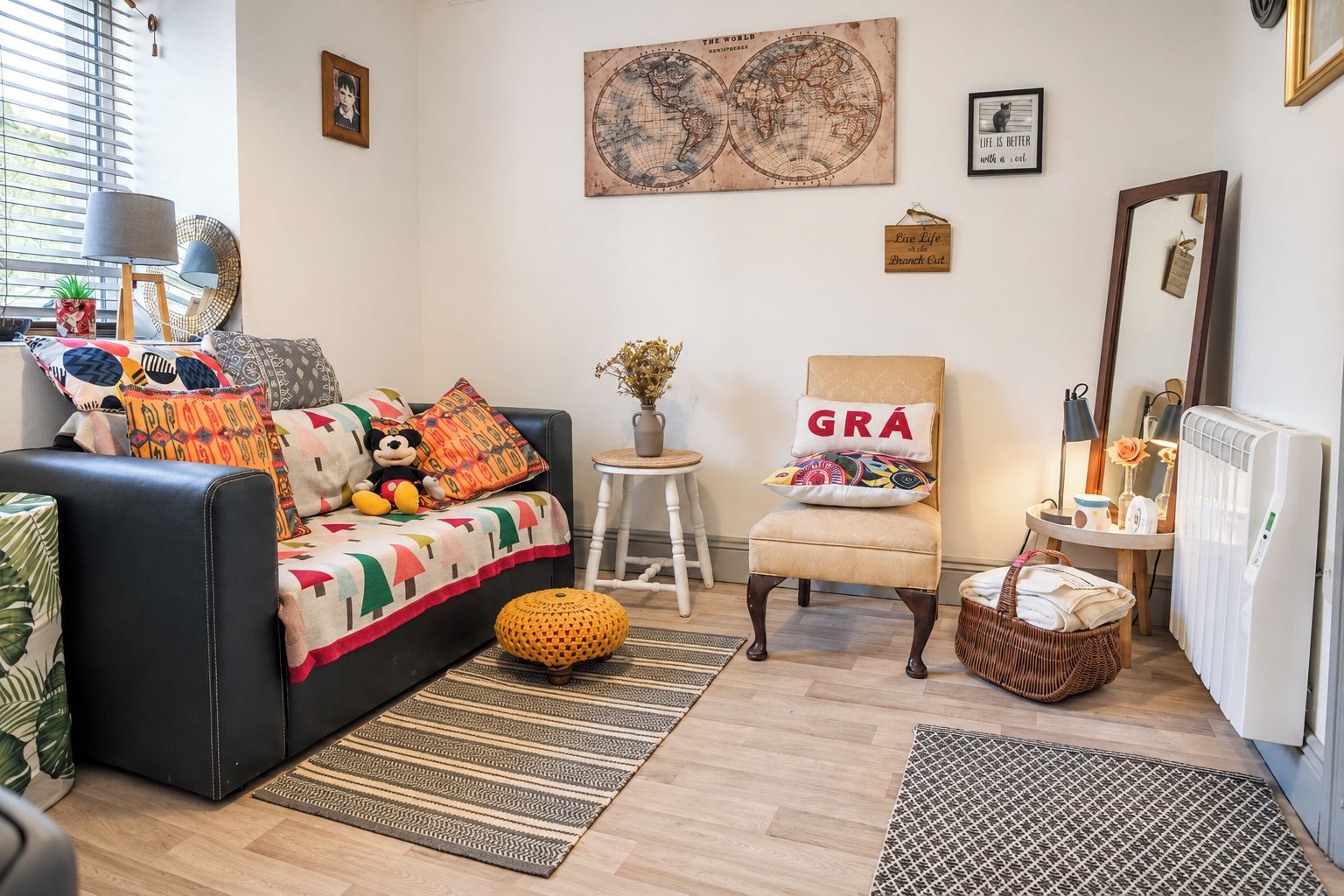 A cozy living room with a black sofa, colorful cushions, a small round table, a chair with a 'GRÁ' pillow, a globe artwork, and a radiator.