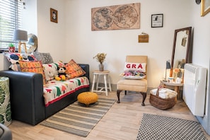 A cozy living room with a black sofa, colorful cushions, a small round table, a chair with a 'GRÁ' pillow, a globe artwork, and a radiator.