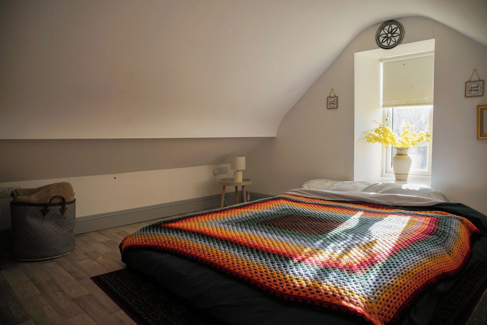 A bedroom with a sloped ceiling, a bed with a colorful blanket, a small bedside table with a lamp, a window with a view of flowers, and a wall-mounted decoration.