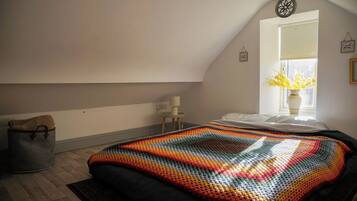 A bedroom with a sloped ceiling, a bed with a colorful blanket, a small bedside table with a lamp, a window with a view of flowers, and a wall-mounted decoration.