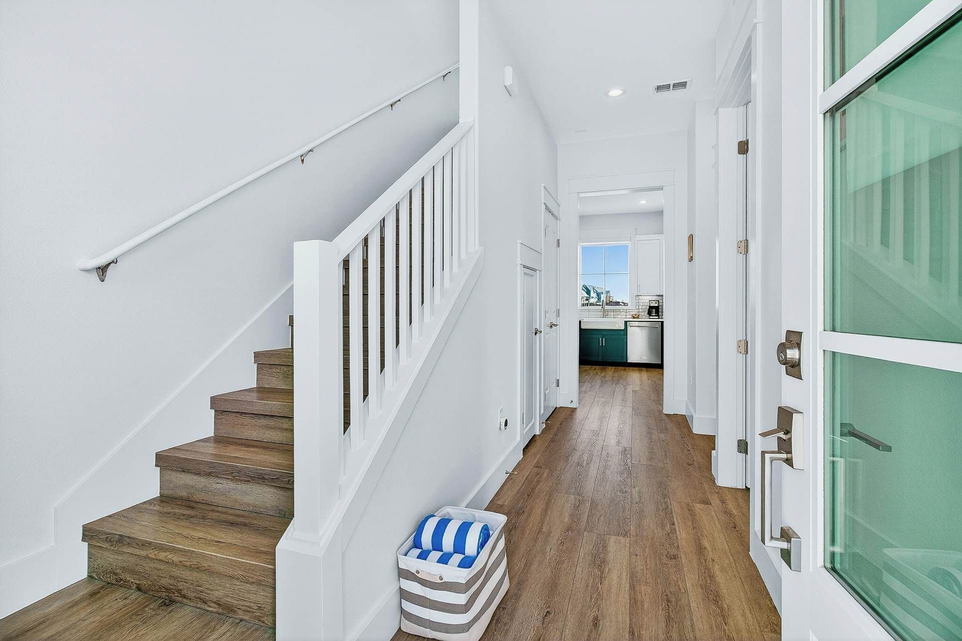 A hallway with a wooden staircase, white walls, and a view into a kitchen area.