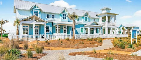 A large, multi-story beachfront property with a white picket fence, paved pathways, and tropical landscaping.