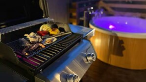 Ein Grill im Freien mit zubereiteten Speisen, ein Whirlpool mit blauer Beleuchtung und eine Holzterrasse.