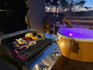 Una parrilla exterior con comida cocinándose, un jacuzzi con iluminación azul y una terraza de madera.