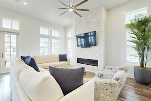 A spacious living room with a high ceiling, a flat-screen TV mounted on a stone wall, a large potted plant, and a fireplace. - Real Good Tan | Golf Views | Steps to Beach (Port Aransas)