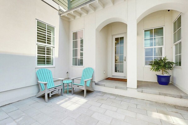 A covered outdoor seating area with two chairs and a table, a potted plant, and a door leading to the interior. - Real Good Tan | Golf Views | Steps to Beach (Port Aransas)
