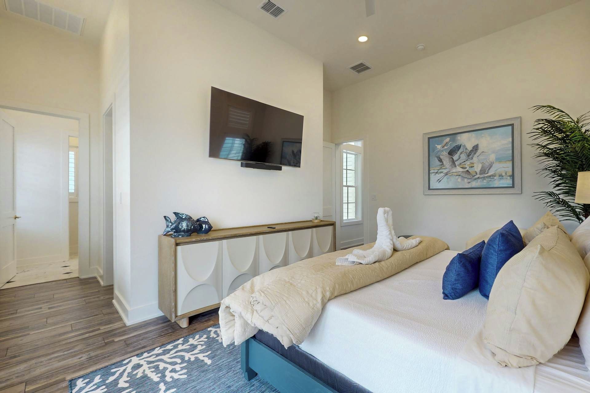 A bedroom with a bed, a TV mounted on the wall, a painting, and a plant.