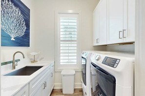 A compact kitchen with a sink, countertop, white cabinets, a window with blinds, and a washing machine. - Real Good Tan | Golf Views | Steps to Beach (Port Aransas)
