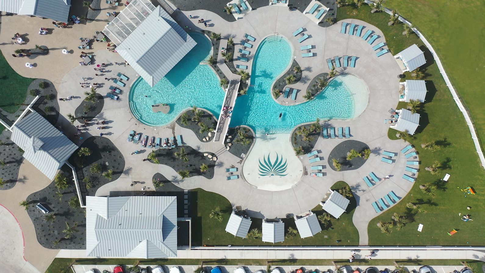 Vista aérea de um resort com uma grande piscina central, várias áreas de descanso e um estacionamento.