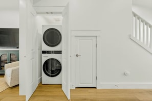 A laundry room with two stacked front-loading washing machines, a white door, and a staircase to the right. - Wave And Sea | Steps to Beach | Resort Amenities (Port Aransas)