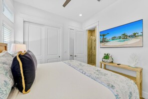 Ein Schlafzimmer mit einem Bett, einem Nachttisch, einem Strandbild und einer Tür zu einem Badezimmer.