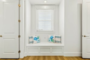 Een kamer met witte wanden, een bank met kussens, een raam met zonwering en een deur met een messing knop.