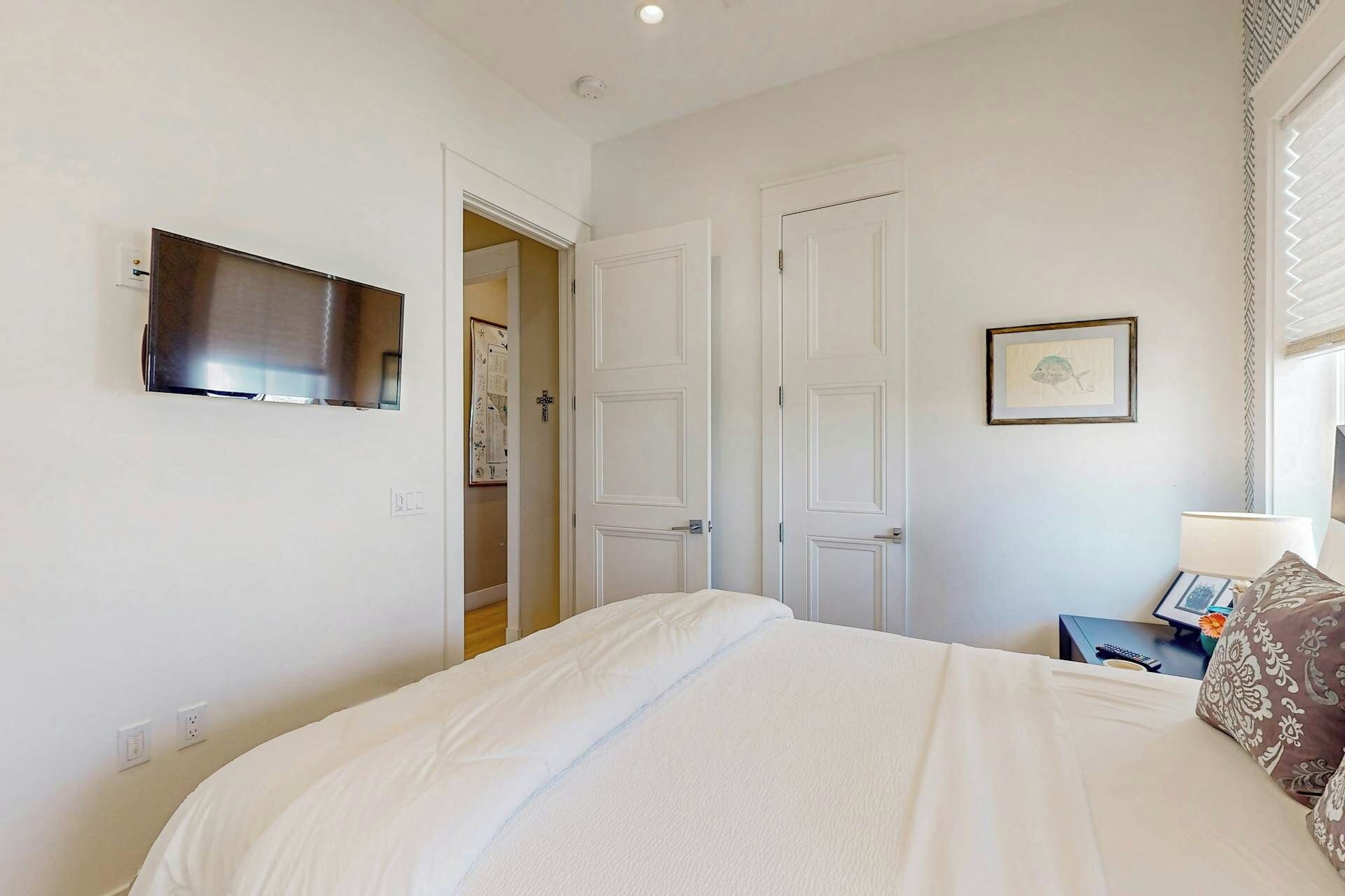 Un dormitorio con cama, televisor montado en la pared, una puerta y un cuadro enmarcado.