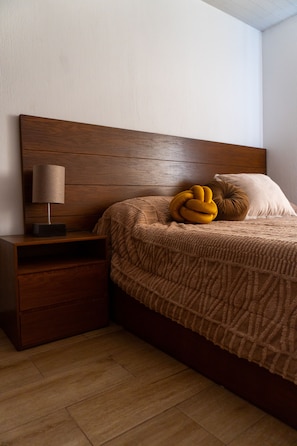 A bedroom with a wooden headboard, a bedside table with a lamp, a bed with a patterned blanket, and two pillows.