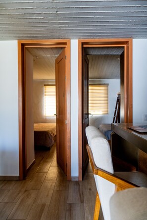 Ein kleines Zimmer mit einem Bett, einem Schreibtisch mit einem Laptop und einem Stuhl. Holztüren führen in einen weiteren Raum.