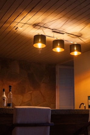 A bar area with wooden ceiling and three pendant lights. There are bottles on the counter and a chair.