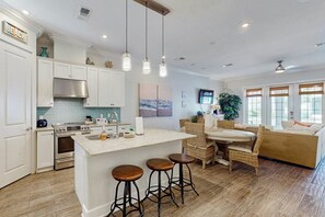 A modern kitchen with white cabinets, a central island with stools, and a dining area with wicker furniture. - Coastal Condo | Private Patio | Resort Amenities (Port Aransas)