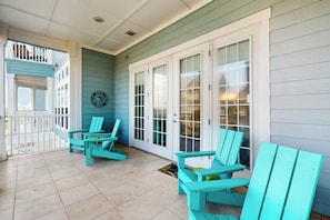 A covered porch with turquoise Adirondack chairs, a clock, and a view of the house. - Resort Views | Covered Balcony | Beach Getaway (Port Aransas)