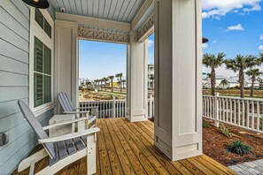 A covered porch with a wooden deck, white railings, and a view of a residential area with palm trees and houses. - Starfish Cove | Steps to Beach | Resort Amenities (Port Aransas)