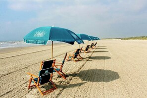 A beach with rows of beach chairs and umbrellas, some with logos, under a clear sky. - Tee - 'N - Sea | Golf Views | Pet Friendly (Port Aransas)