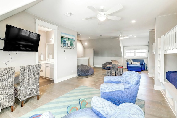 A spacious living area with a flat-screen TV, seating, a ceiling fan, and a large window. - Top Of The Shoreline | Golf Views | Steps to Beach (Port Aransas)