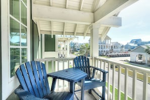 A covered porch with two chairs and a table, overlooking a residential area with houses and a street. - Top Of The Shoreline | Golf Views | Steps to Beach (Port Aransas)