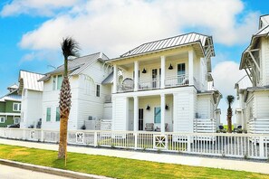 A two-story white building with a balcony, a white picket fence, and a palm tree in the foreground. - Vida Del Mar | Steps to Beach | Resort Amenities (Port Aransas)