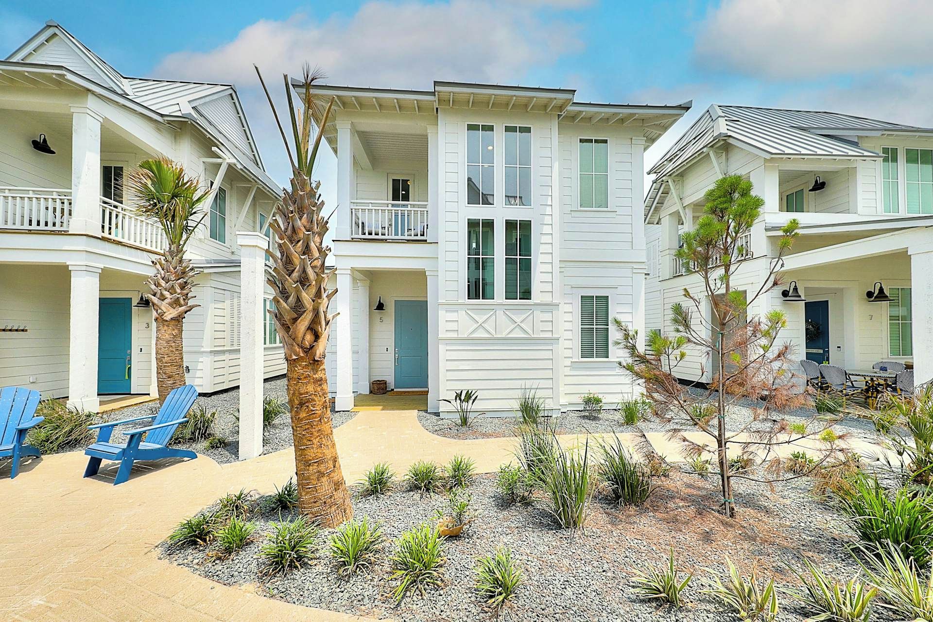 A beachfront property with multiple-story buildings, a covered porch, and a sandy patio area with blue chairs.