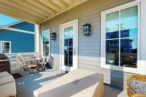 A covered outdoor area with a table and chairs, a bench, and a view of a building through large windows. - We Be Beachin' | Golf Views | Pet Friendly (Port Aransas)
