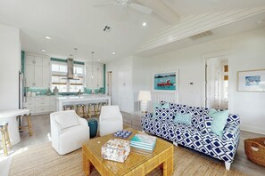 Un salon moderne avec un canapé blanc, une table basse et un coin cuisine doté de meubles blancs et d’un plan de travail bleu.
