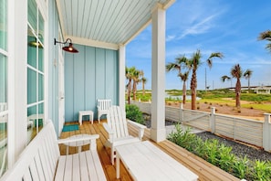 A covered outdoor area with white chairs and a table, overlooking a landscaped area with palm trees and a view of the beach. - Palmilla Beach Escape | Steps to Beach (Port Aransas)