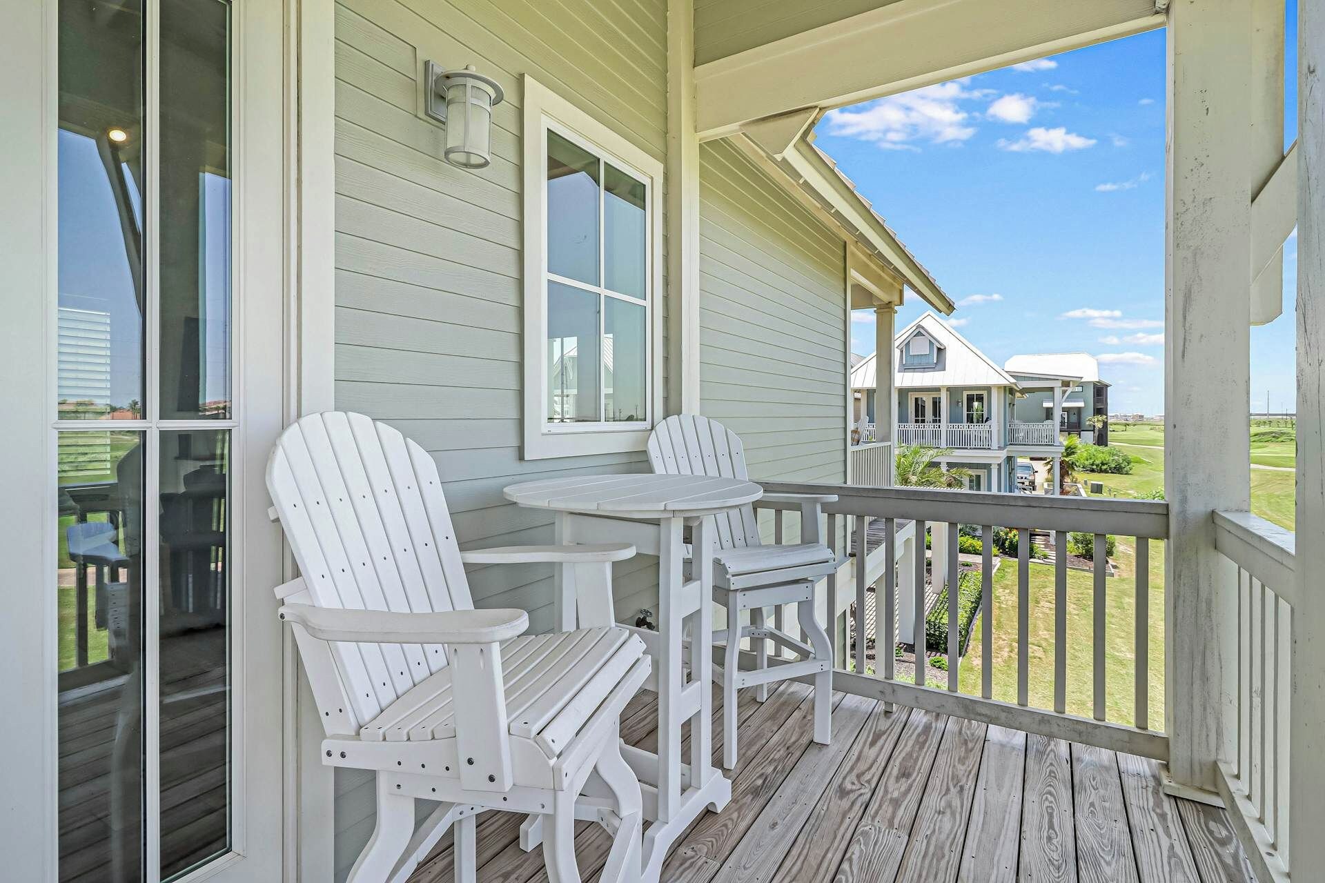 A covered porch with two Adirondack chairs and a small table, overlooking a grassy area with a house in the background.