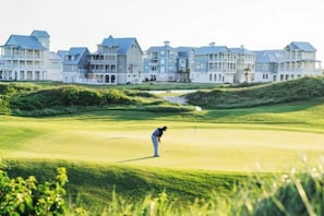 Een golfbaan met een persoon in een witte outfit die zich klaarmaakt om te putten, omringd door een clubhuis met meerdere gebouwen.