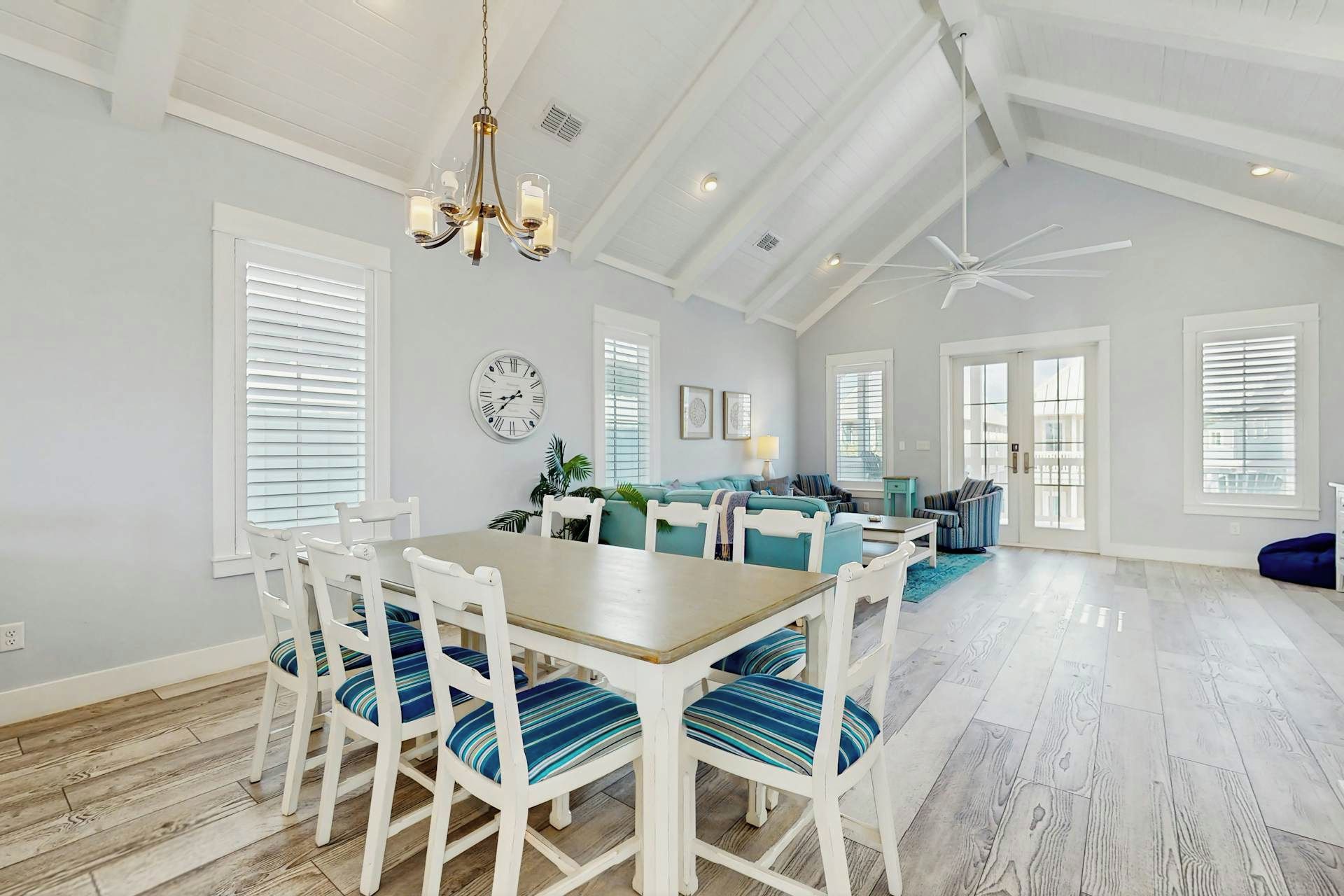 A spacious dining area with a wooden table and white chairs, a ceiling fan, and a large window with blinds.