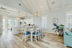 A spacious kitchen with white cabinetry, a dining area with white chairs, and a large clock on the wall. - Paradise Found | Steps to Beach | Resort Amenities (Port Aransas)