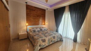 A bedroom with a wooden headboard, a bed with patterned bedding, a nightstand, and a large window with curtains. - Poolside One-Bedroom Retreat @ Villa Troyanda in Grace Bay (Providenciales)