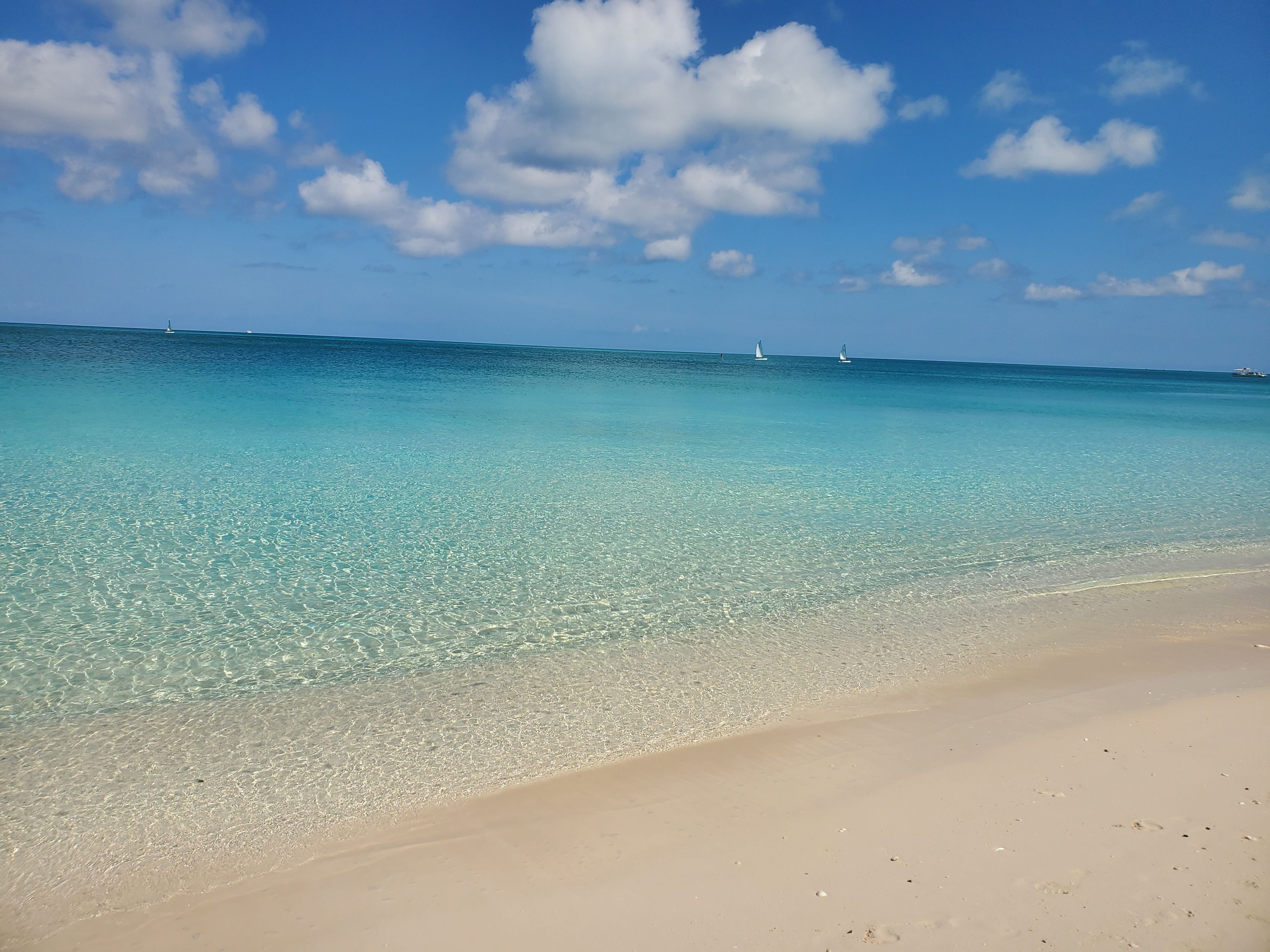 A beach with clear turquoise water, a sandy shore, and a few sailboats in the distance.